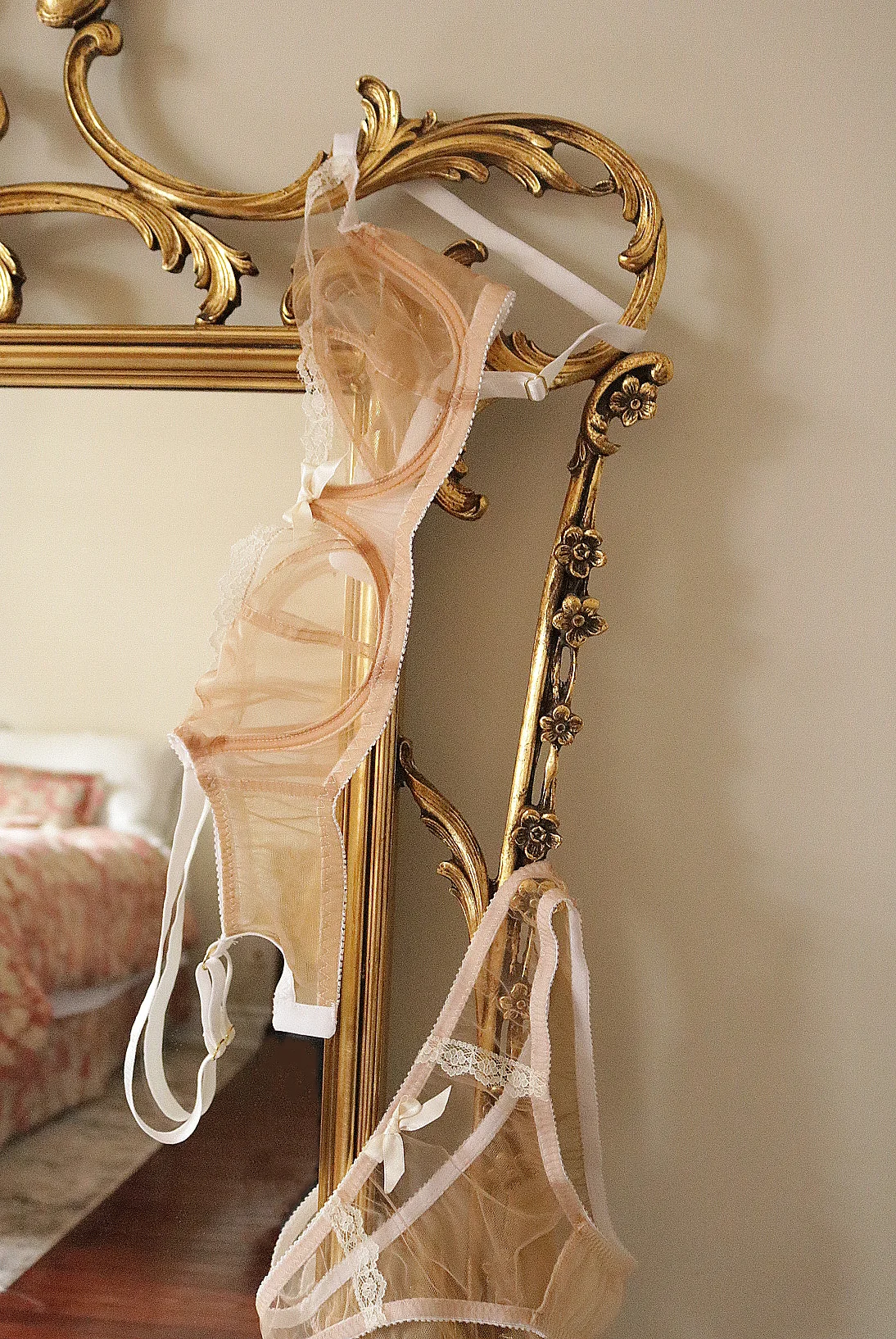 Sheer peach bra and panty hanging on an ornate gold-framed mirror in a bedroom with floral bedding