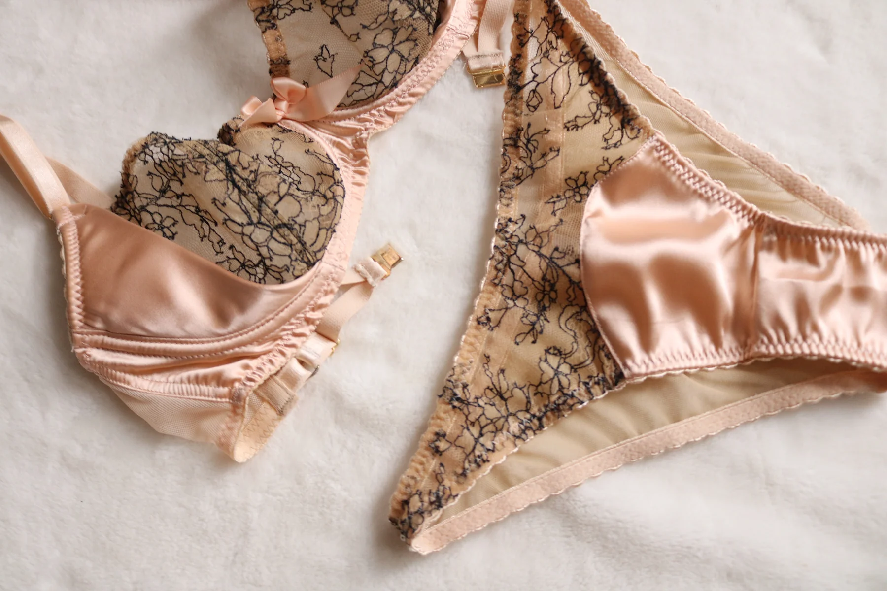 Peach satin bra and underwear with black floral lace overlay, laid flat on a soft white textured surface