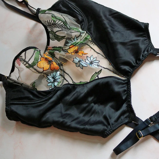 Flat lay of black satin lingerie piece with sheer floral embroidered panel in orange, blue, and green, laid on a light surface