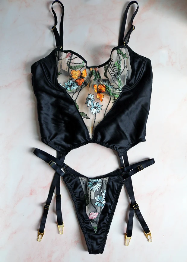 Black satin bodysuit with colorful floral mesh panels and garter straps, laid flat on a marbled surface